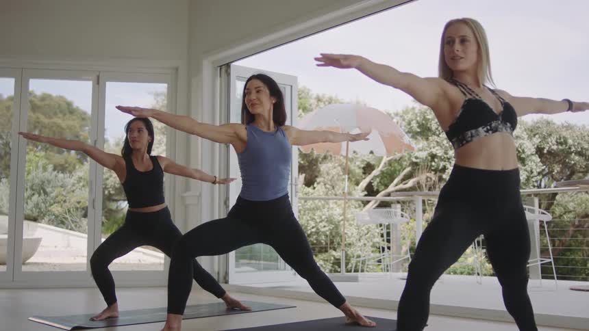 Three diverse women transition from a side stretch to a Warrior II pose during a yoga class in a bright, airy studio