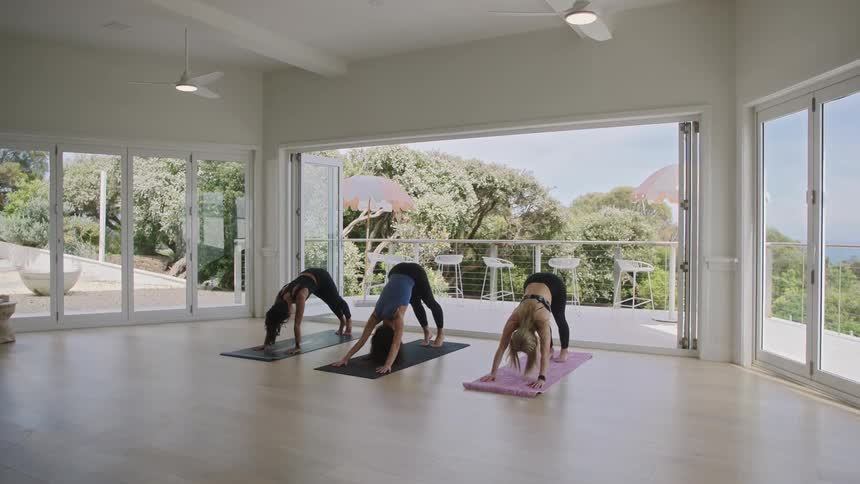 Three women hold the downward-facing dog yoga pose in a bright, modern studio with large open doors looking out onto a lush balcony and the ocean
