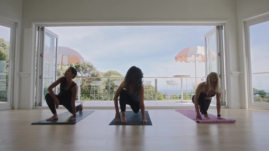 Three women gracefully transition through yoga poses in a beautiful, open-air studio with an ocean backdrop