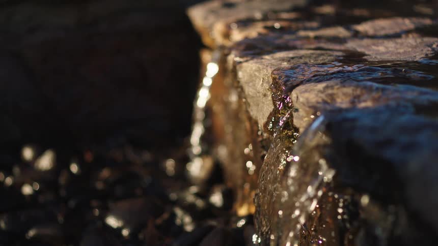 A tranquil close-up, slow-motion shot of water trickling over a stone fountain ledge, illuminated by the warm glow of sunlight