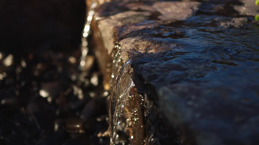 A slow-motion close-up of a tranquil man-made waterfall cascading over flat stones into a bed of pebbles