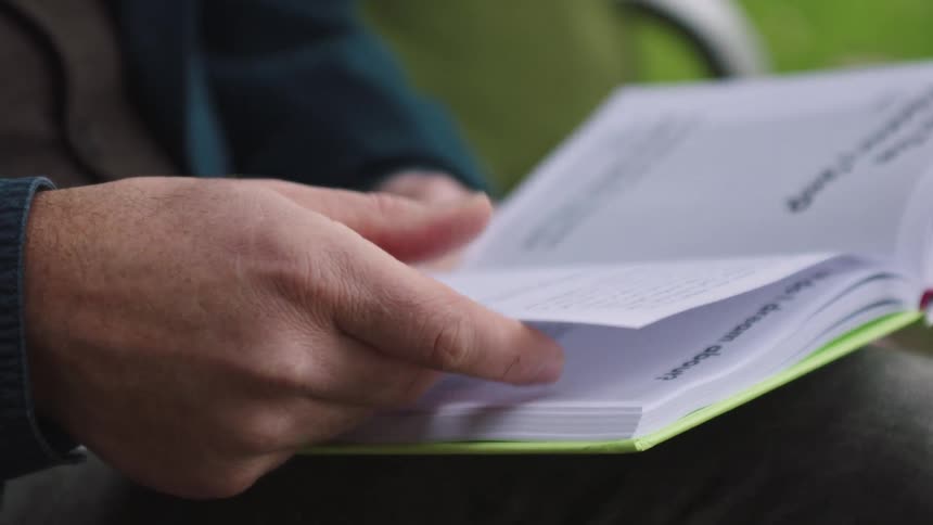 A close-up slow-motion shot of a man's hands slowly turning the pages of a book while sitting on a park bench