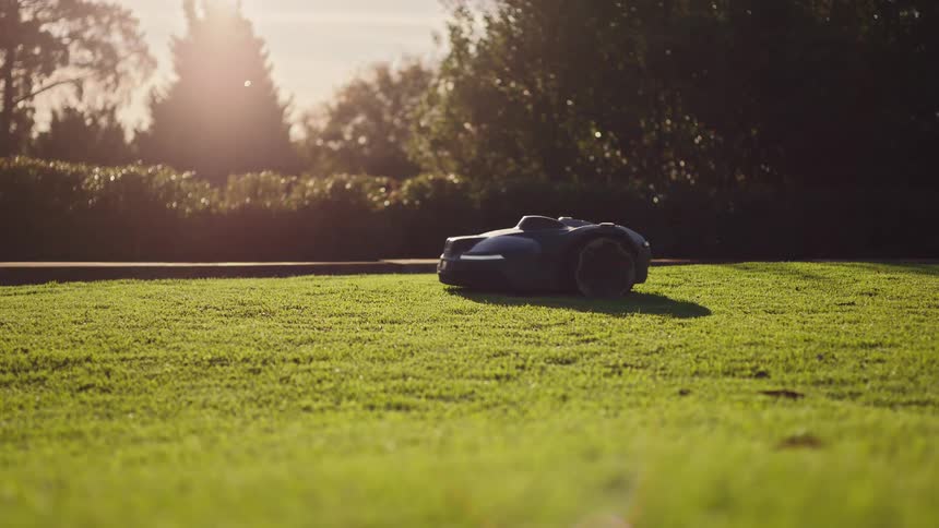 A robotic lawn mower trims a lush green lawn as the sun sets, casting a warm golden light