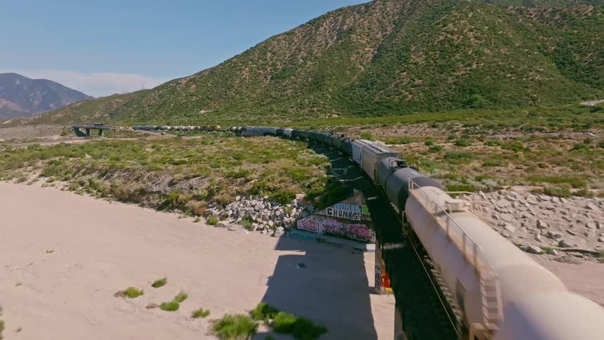An aerial view follows a freight train with tanker cars as it snakes through a vast, arid valley at the base of green hills