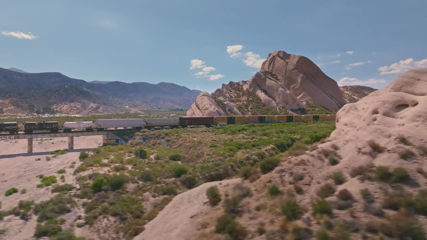 An aerial shot tracks a long freight train as it crosses a bridge in a rugged desert landscape with unique rock formations
