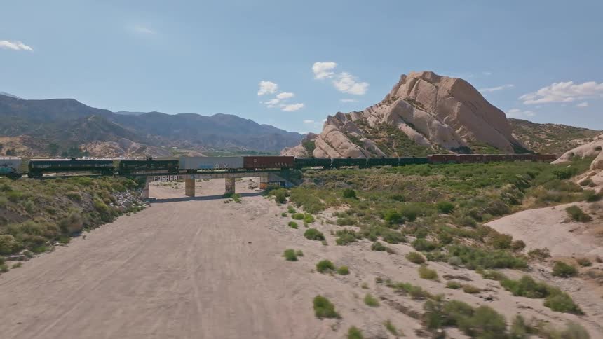 A cinematic aerial shot pans to reveal a freight train crossing a bridge amidst the dramatic rock formations of a desert landscape