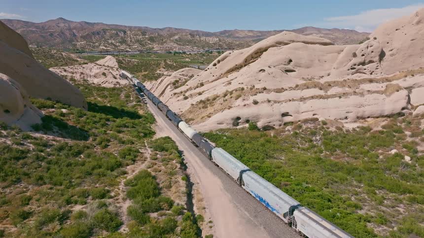An aerial view shows a long freight train moving slowly through a scenic, rocky desert canyon on a sunny day