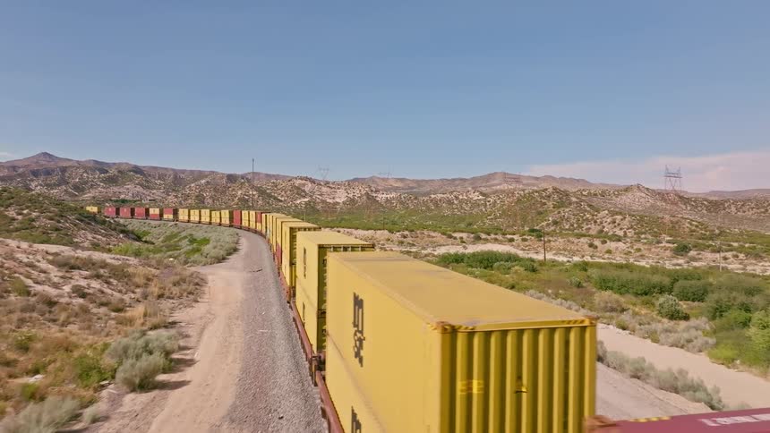 An aerial view of a long freight train carrying shipping containers as it winds through a vast desert landscape on a sunny day