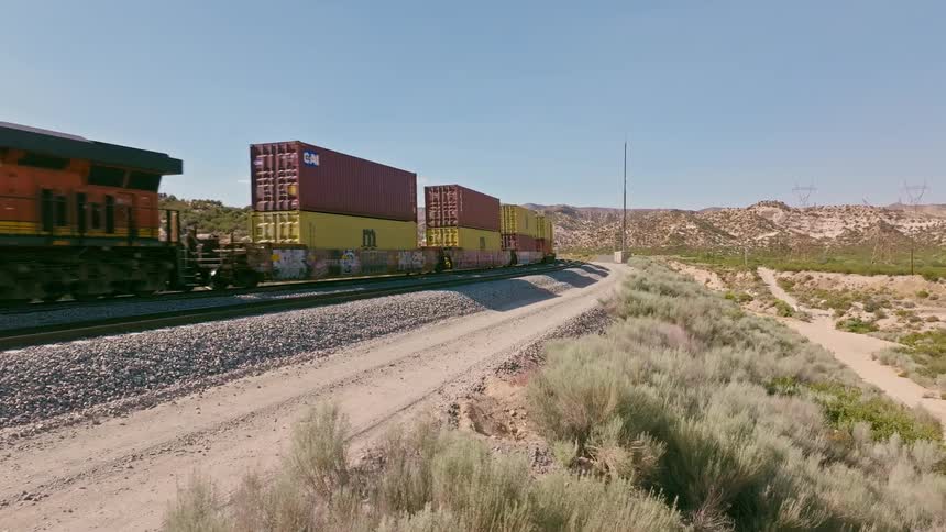 A freight train with double-stacked shipping containers rolls along the tracks, receding into the distance through a vast, arid desert landscape