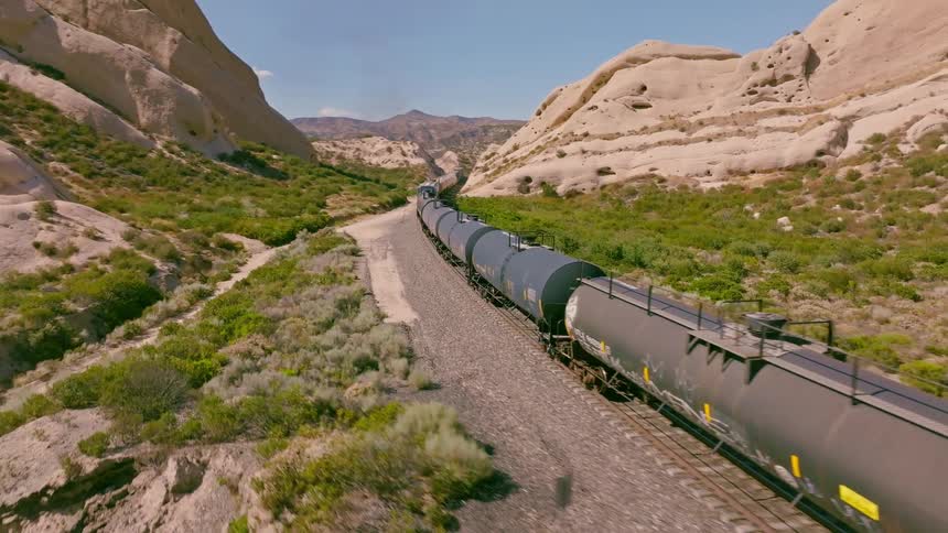An aerial view of a long freight train with tanker cars traveling over a bridge and through a vast desert canyon with unique rock formations