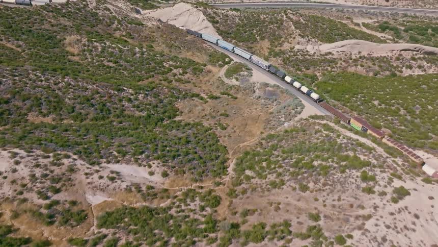 An aerial drone shot flies over a smooth, sandy rock formation, revealing a freight train moving through a dry, hilly landscape