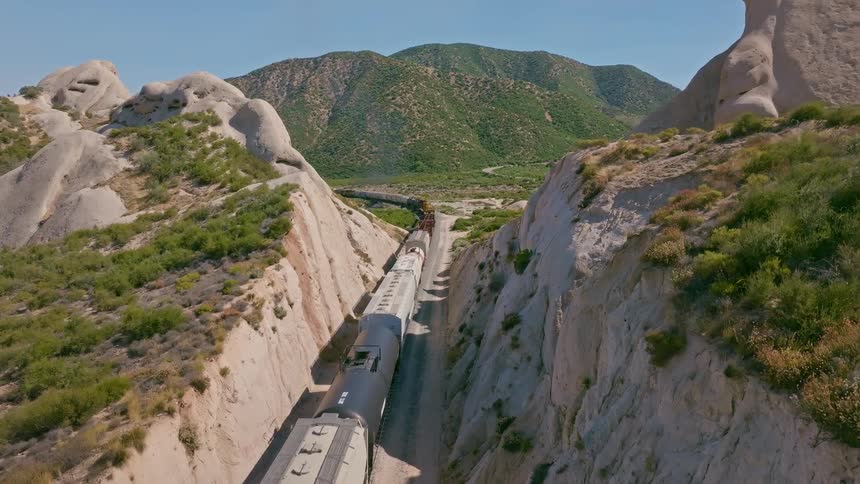 An aerial view captures a long freight train slowly passing through a dramatic, narrow rock canyon in a sun-drenched mountain landscape