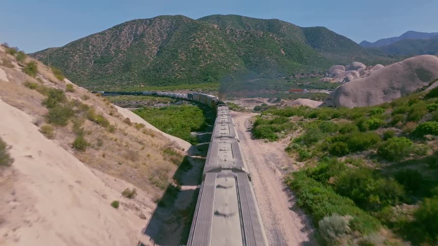 A dramatic aerial view follows a long freight train as it travels through a narrow, rocky canyon in a vast desert mountain landscape