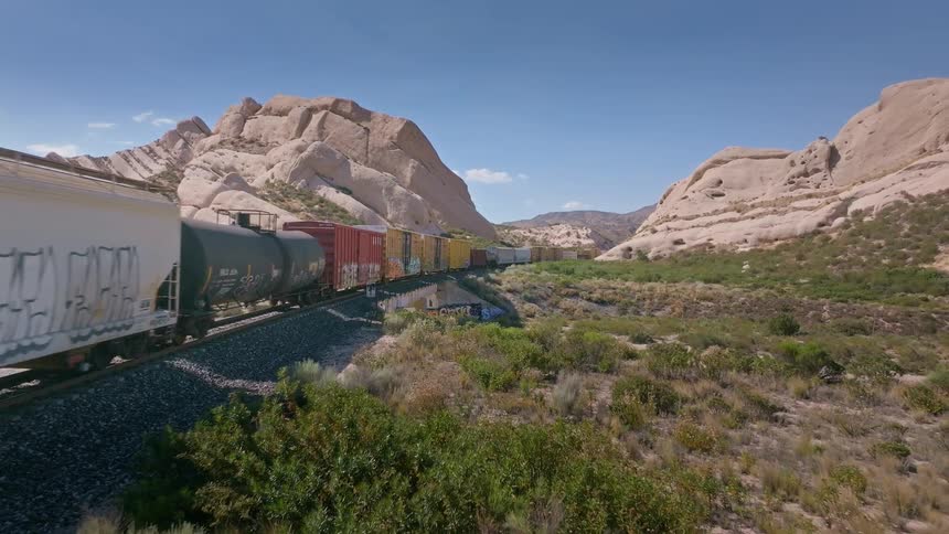 A long, colorful freight train with graffiti art travels through a rocky desert canyon landscape on a bright sunny day
