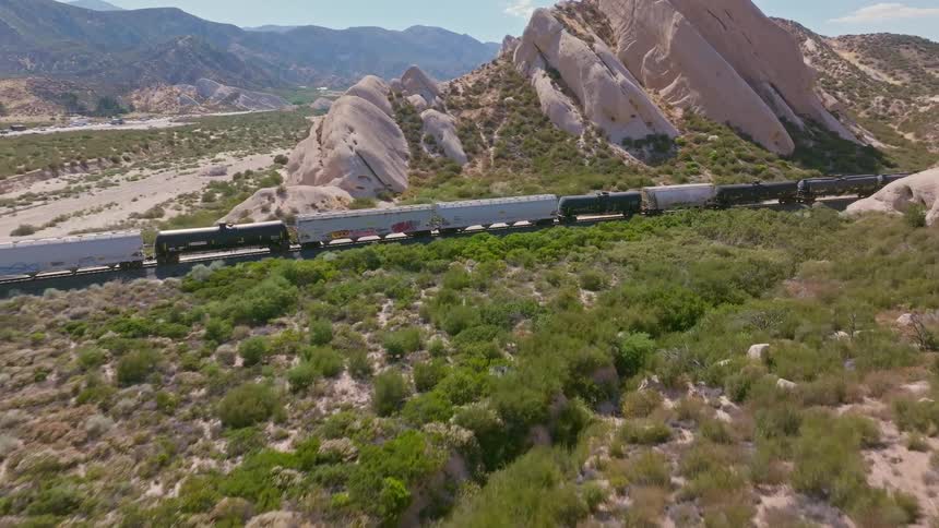 An aerial drone flies over a rugged desert hill, revealing a long freight train traveling alongside dramatic rock formations
