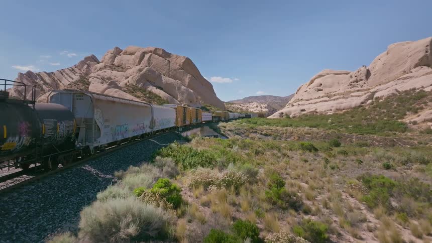An aerial shot follows a long freight train covered in graffiti as it travels along a railway through a vast, rocky desert canyon on a sunny day