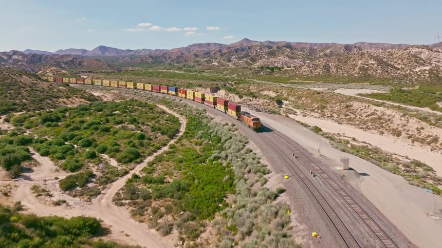 An aerial shot reveals a long, colorful freight train curving through a vast, arid desert landscape under a clear blue sky