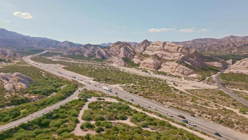 An expansive aerial view reveals traffic flowing on a highway alongside railroad tracks through a vast desert landscape with unique rock formations