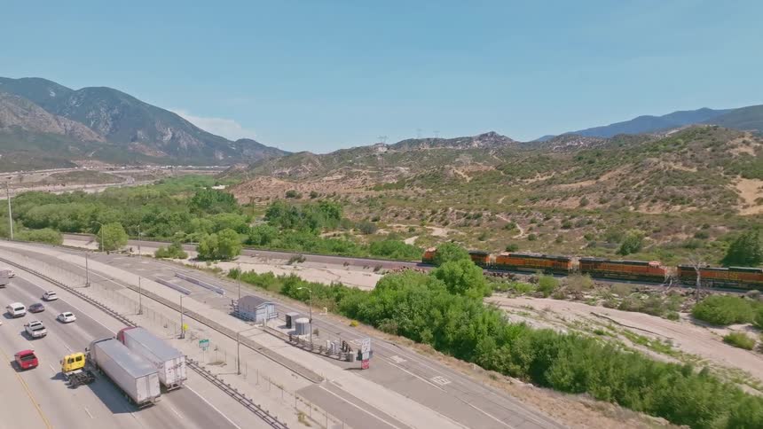 Aerial view of a bustling highway and a passing freight train running parallel through a rugged mountain valley on a sunny day