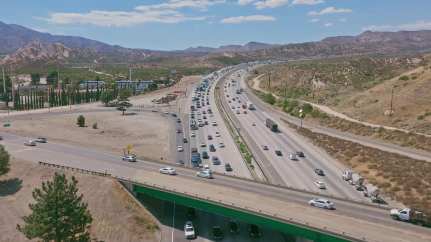 Drone flies over a major multi-lane highway with heavy traffic congestion flowing through a dry, hilly landscape on a sunny day