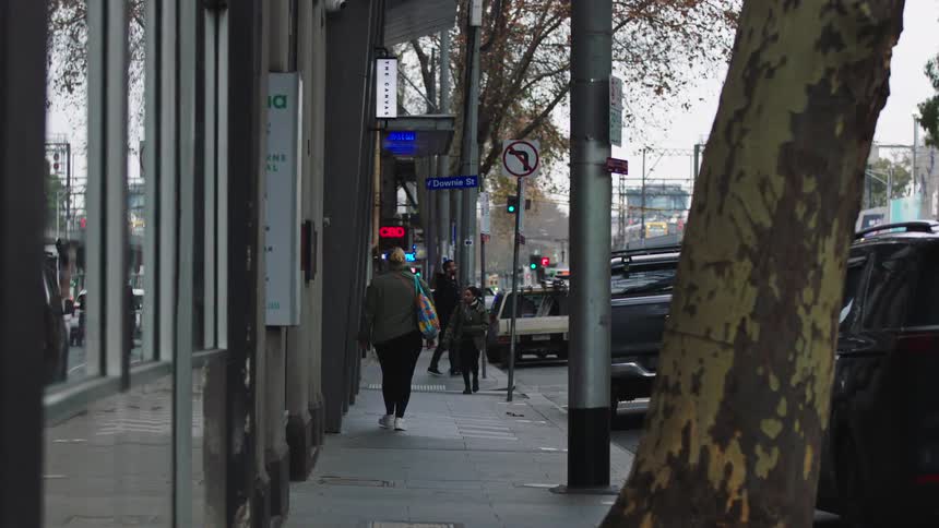 A person wearing a green jacket and carrying a colorful bag walks down a city sidewalk in slow motion