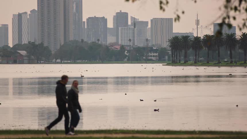 An out-of-focus couple walks in slow motion along a tranquil city lake, with ducks swimming and a hazy skyline in the background