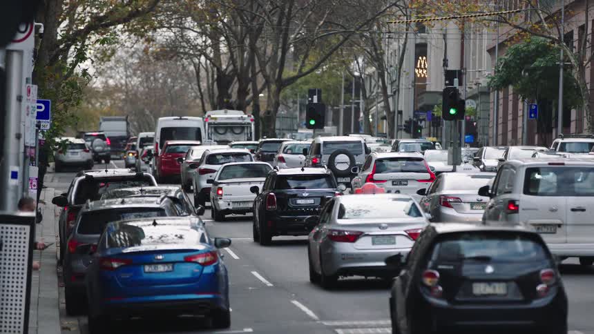 Slow-moving traffic creates congestion on a busy city street lined with trees and buildings