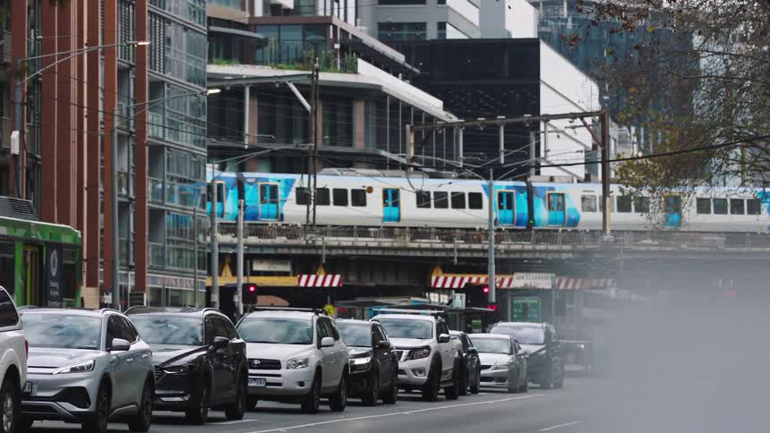 A slow-motion view of a bustling city street shows heavy traffic as a metro train crosses an elevated bridge