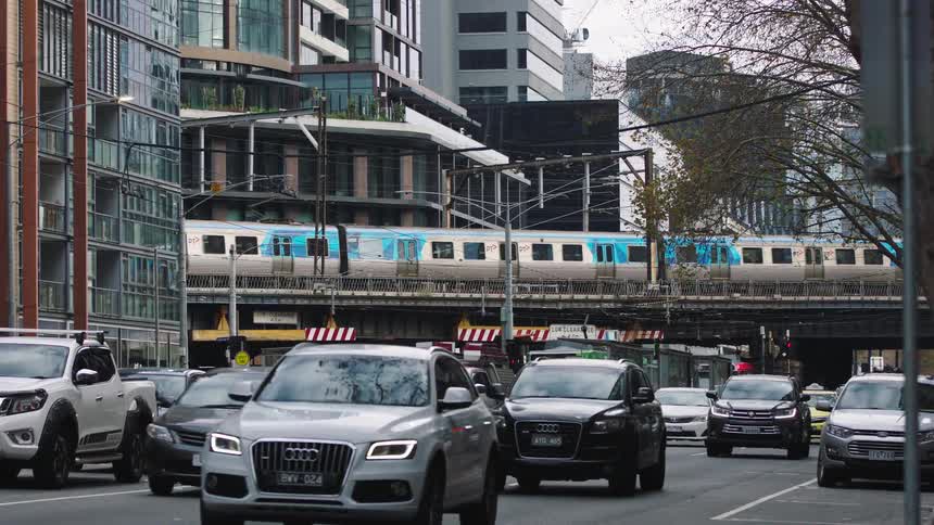 A slow-motion shot of a bustling city street with cars in traffic as an elevated train passes overhead on a bridge