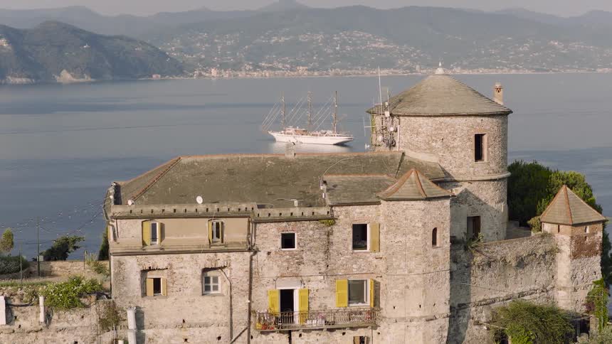 An old stone castle with yellow shutters sits on a cliffside overlooking the water as a beautiful multi-masted sailboat glides by