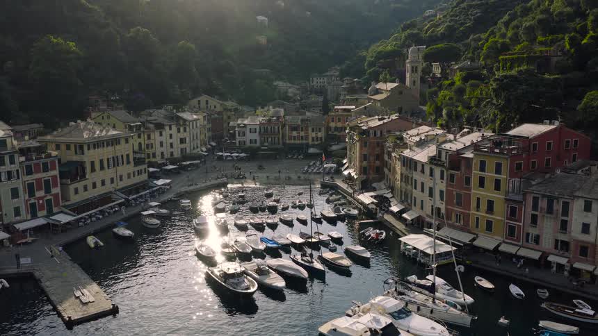 Aerial drone footage gracefully approaches the sun-drenched harbor of Portofino, Italy, showcasing its iconic colorful waterfront and fleet of luxury yachts and traditional boats