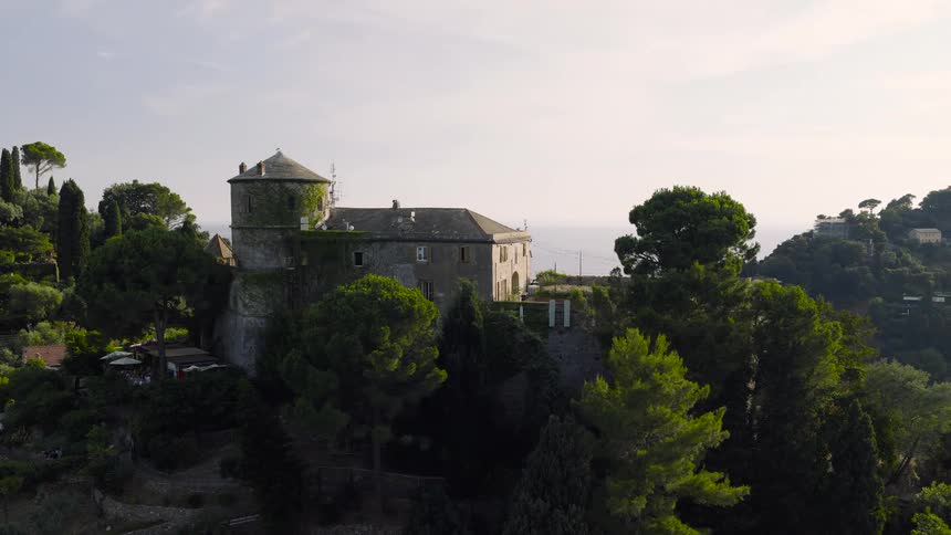 Aerial orbit of the historic Castello Brown, a beautiful stone fortress surrounded by lush trees overlooking the Mediterranean Sea in Portofino, Italy