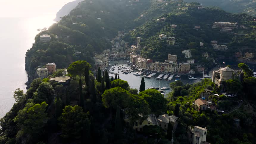 An aerial drone shot flies over the lush green cliffs of the Italian Riviera, revealing the picturesque harbor of Portofino with its colorful buildings and moored yachts