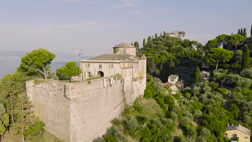 An aerial view reveals the stunning Castello Brown in Portofino, perched on a verdant hill overlooking the Ligurian Sea as the camera slowly orbits