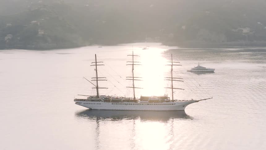 A majestic tall ship is anchored in a tranquil bay off a scenic coastal town as the morning sun breaks through the haze
