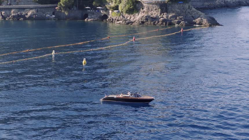 A classic wooden speedboat with passengers rests on the calm blue waters near a rocky European coastline
