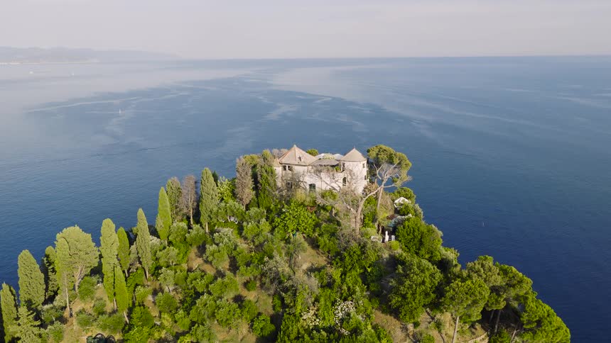 An aerial view slowly advances towards a castle and lighthouse on the lush Portofino peninsula, surrounded by the beautiful blue Ligurian Sea