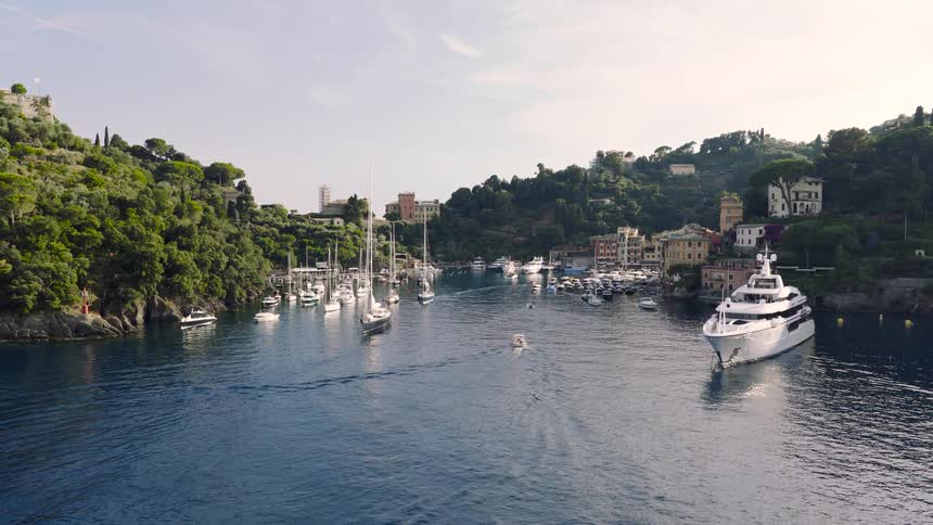 A serene aerial approach to the picturesque harbor of Portofino, where luxury yachts and sailboats are moored along the lush Italian Riviera coastline