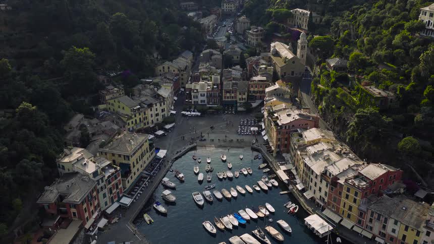 An elegant aerial view slowly pulls back to reveal the stunning and picturesque harbor of Portofino, Italy, filled with boats and surrounded by lush hillsides