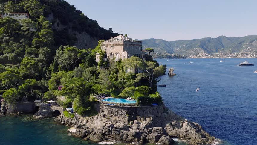 A magnificent historic villa with a scenic swimming pool stands on a lush, rocky cliff above the blue waters of the Italian Riviera