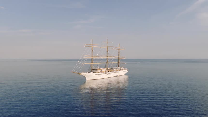 A majestic white three-masted tall ship is anchored in the calm, deep blue sea as the aerial camera slowly pulls back for a wide view