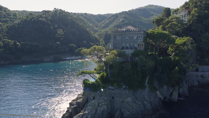 Aerial view of a stunning historic villa perched on a rocky cliff overlooking the sparkling sea and lush green coastline of Portofino, Italy