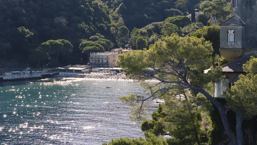 A slow pan from a historic villa on a cliffside reveals a sun-drenched bay with a busy beach on the picturesque Italian coastline