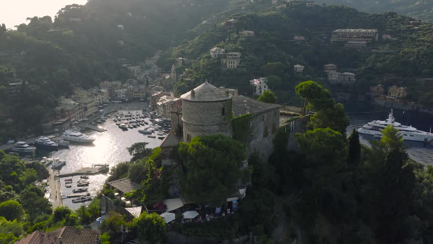 An ascending aerial shot reveals the picturesque harbor of Portofino, Italy, with the historic Castello Brown overlooking the village and marina