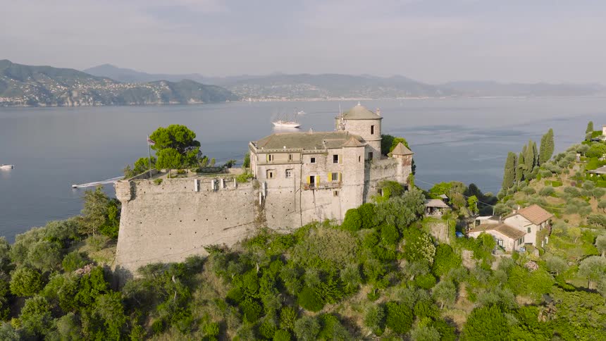 Slowly pulling back aerial shot of the historic Castello Brown fortress on a green cliff in Portofino, Italy, with boats on the sea and mountains in the distance