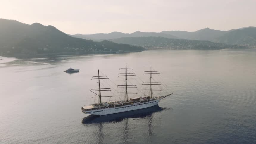 An aerial drone flies over a majestic tall ship anchored in a tranquil bay with a mountainous coastline in the background during a hazy sunrise