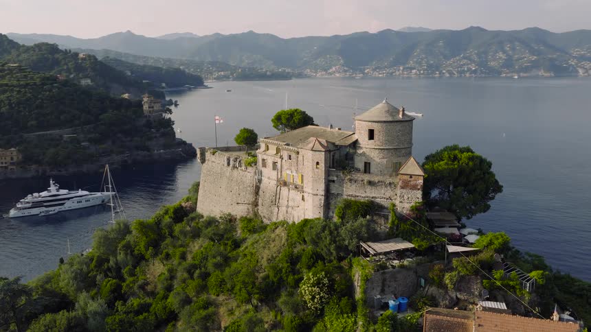 An aerial view orbits the historic Castello Brown perched on a lush cliffside overlooking the idyllic harbor and coastline of Portofino, Italy