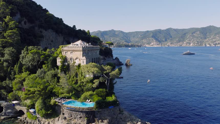 A stunning aerial view flies towards a luxurious historic castle with a pool on the beautiful Portofino coastline in Italy