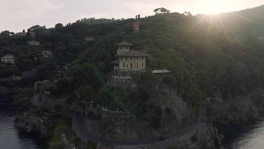An aerial shot slowly reveals a stunning Italian villa built into a lush, green cliffside overlooking the sea as the sun begins to set
