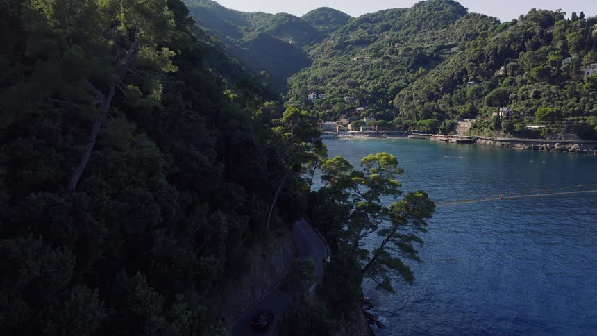 Aerial view of a scenic coastal road winding along a lush, green mountainside next to the deep blue Mediterranean Sea